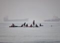 Varias personas se congregan en tablas de remo en aguas poco profundas al tiempo que buques de carga y de servicio permanecen anclados en el estrecho de Ormuz
