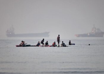 Varias personas se congregan en tablas de remo en aguas poco profundas al tiempo que buques de carga y de servicio permanecen anclados en el estrecho de Ormuz