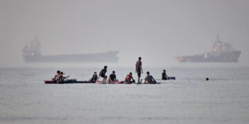 Varias personas se congregan en tablas de remo en aguas poco profundas al tiempo que buques de carga y de servicio permanecen anclados en el estrecho de Ormuz