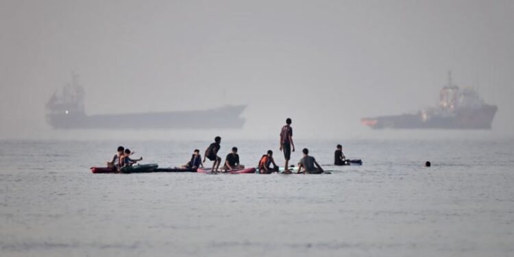 Varias personas se congregan en tablas de remo en aguas poco profundas al tiempo que buques de carga y de servicio permanecen anclados en el estrecho de Ormuz